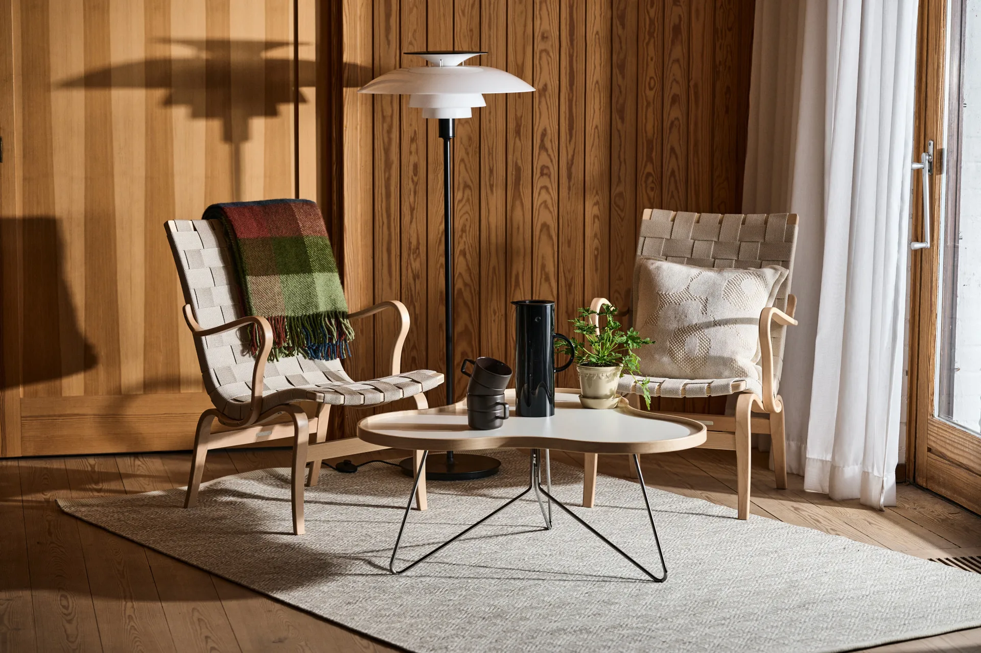 Mysigt rum med två ljusa trästolar med vävda säten, ett vitt soffbord och en golvlampa.