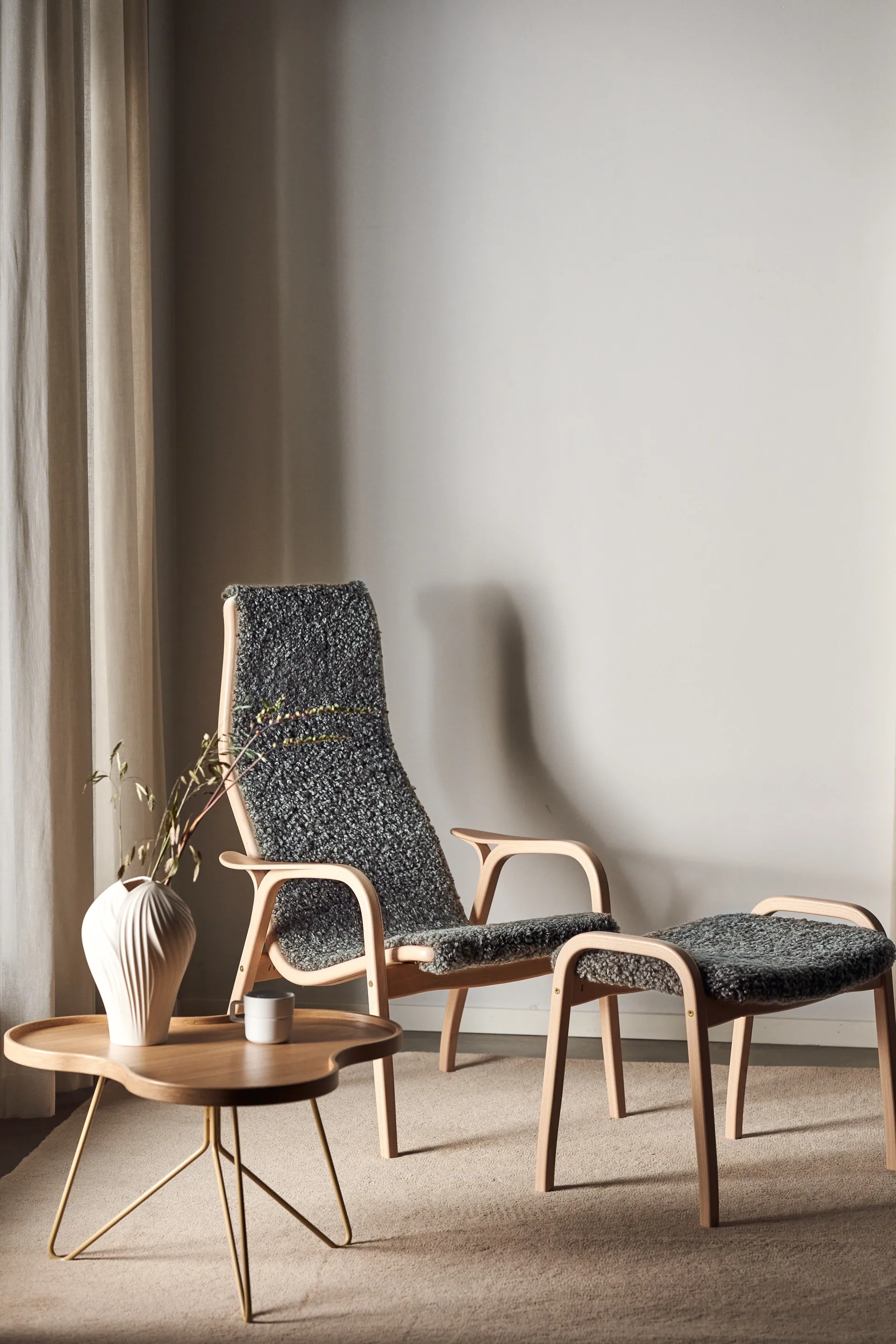 En loungestol i ljust trä med matchande pall och mörkgrå texturerad klädsel, bredvid ett litet soffbord i trä med guldben, vas och mugg.