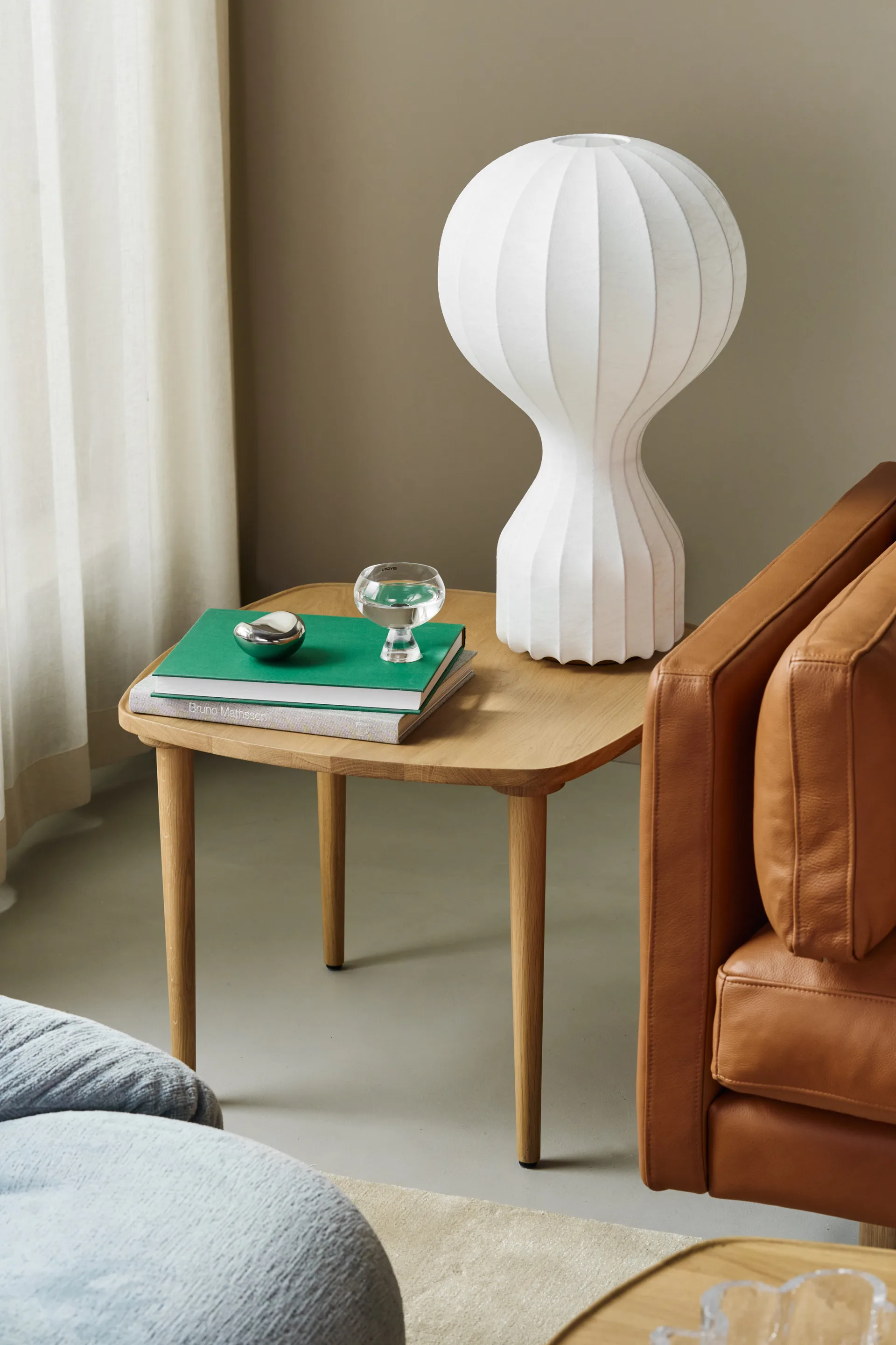 Litet träbord med vit räfflad lampa, gröna böcker, ett vattenglas och ett silverföremål bredvid en brun skinnfåtölj.