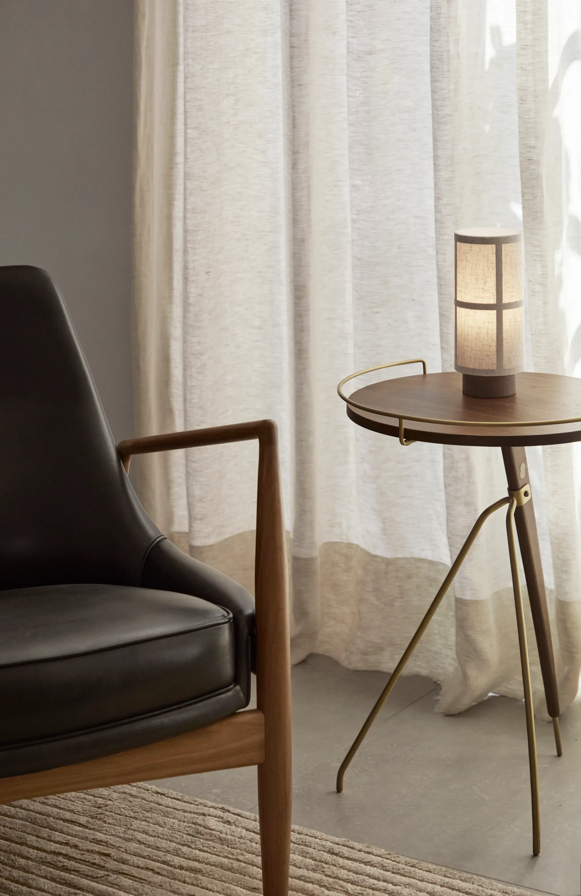Audo Copenhagen svart läderfåtölj, runt sidobord i trä med tänd cylinderformad lampa och skira gardiner.