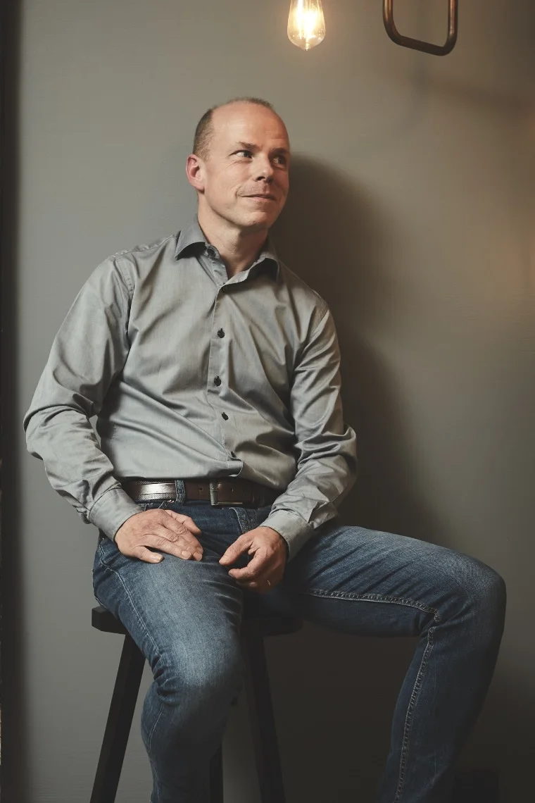 En man i grå skjorta och jeans sitter på en pall och tittar åt höger. En glödlampa hänger ovanför.