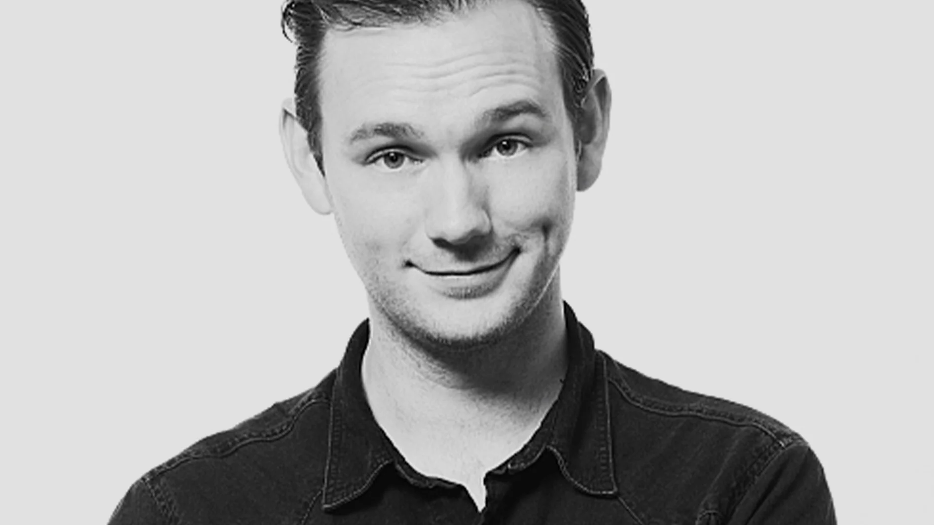Svartvitt porträtt av en leende ung man med kort mörkt hår och smilgrop, iklädd en mörk skjorta.