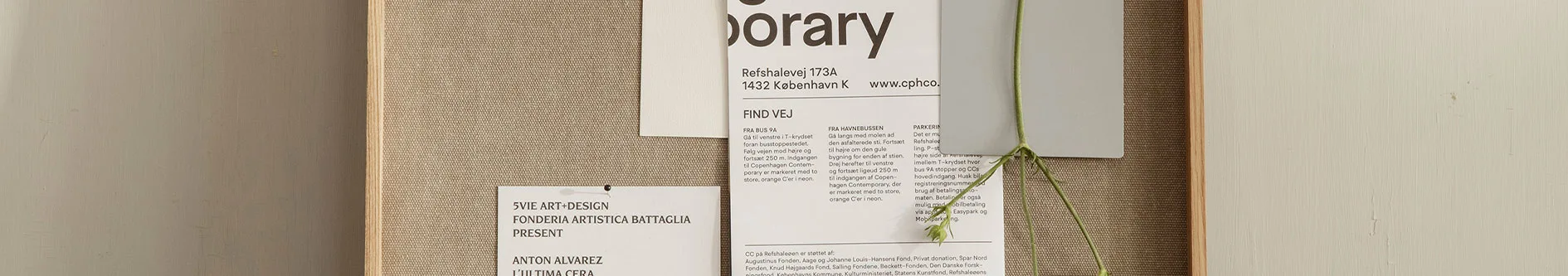 En ljusbrun anslagstavla med vävd textur, inramad i trä. Två vita papper är fästa med häftstift, och en grön växtstjälk hänger över ett av dem.