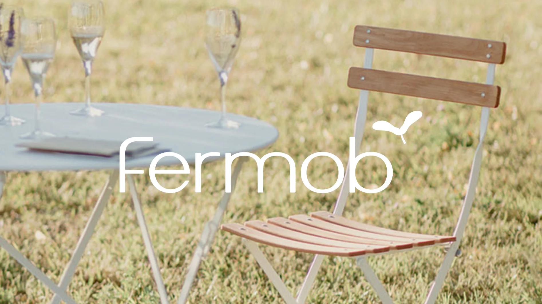 Fermob utemöbler: ett runt bord och en hopfällbar stol i trä och metall på gräs. Tre champagneglas.