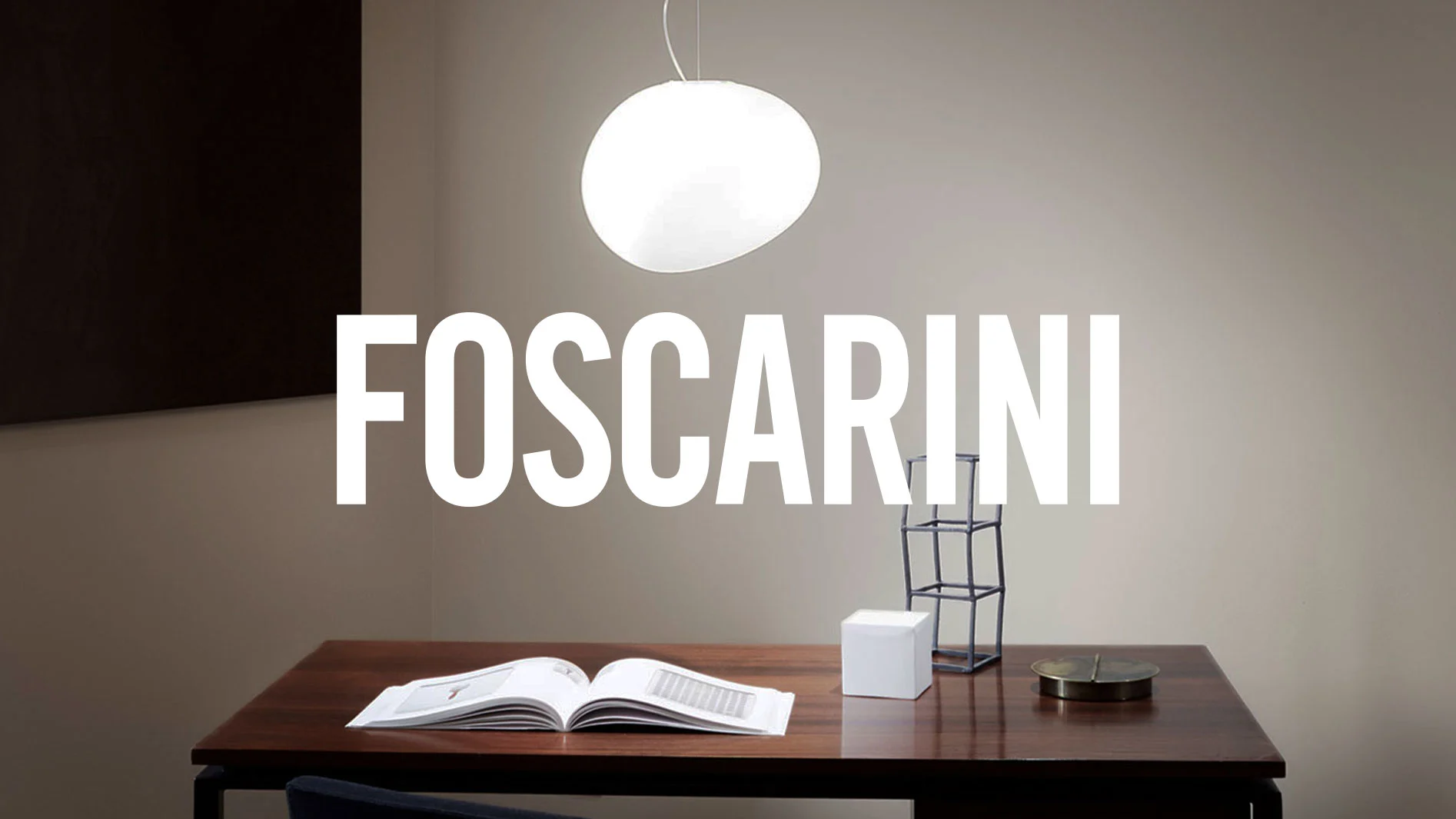"FOSCARINI"-text över ett modernt skrivbord med en upplyst vit taklampa och dekorativa föremål.