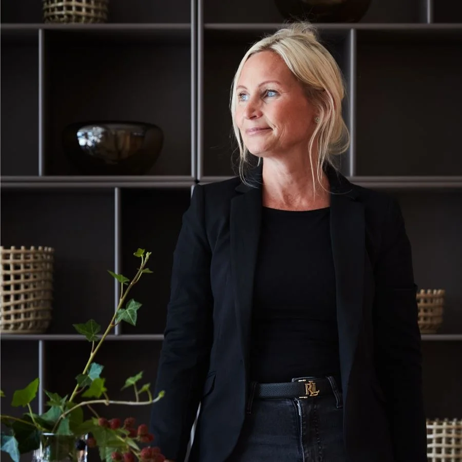 Blond kvinna i svart blazer, mörka jeans med guldfärgat RL-bältesspänne, tittar åt höger. Mörka hyllor i bakgrunden, växt i förgrunden.