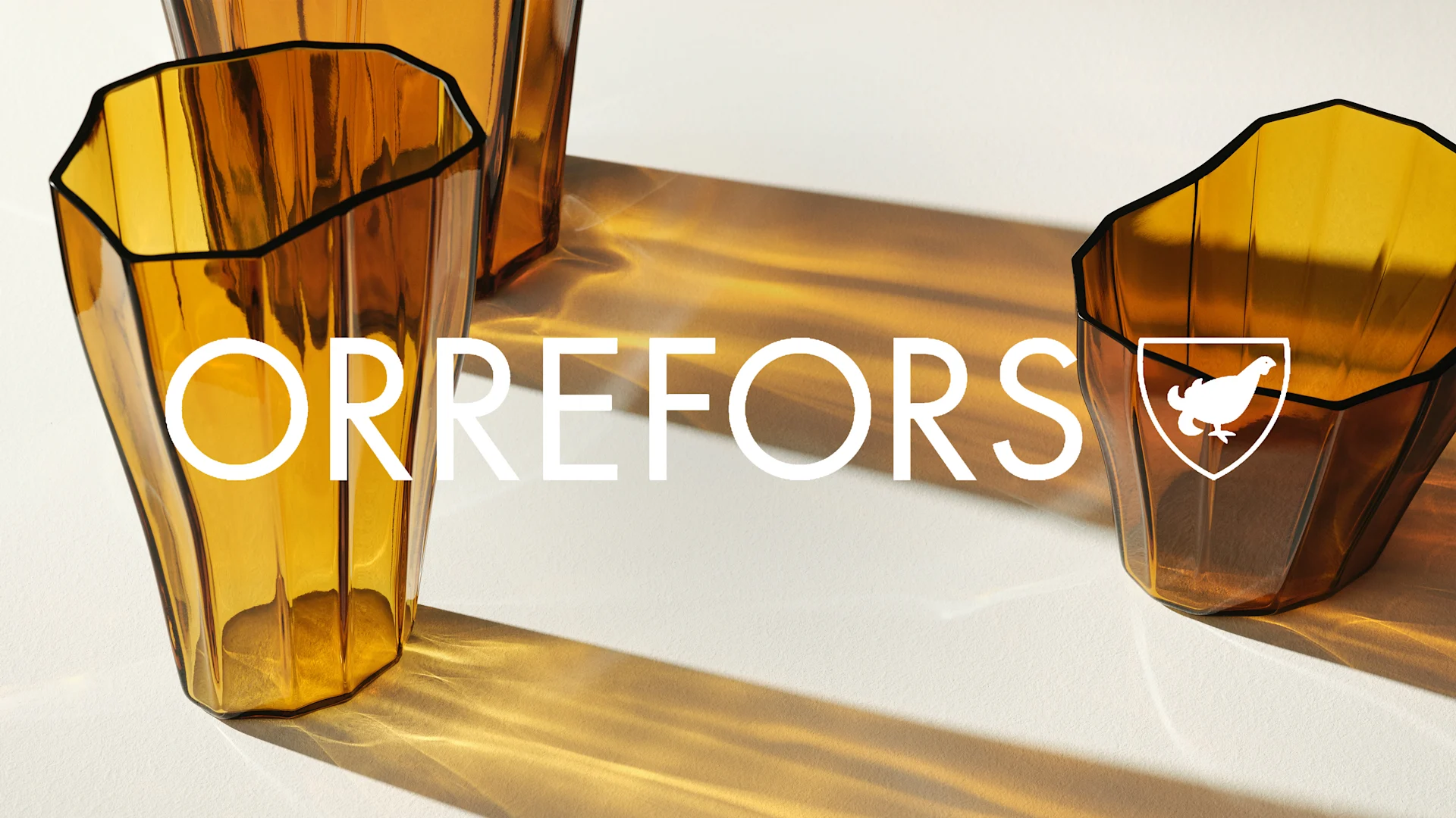 Två bärnstensfärgade, fasetterade glas med Orrefors-logotyp och text, med ljus som skapar mönster.