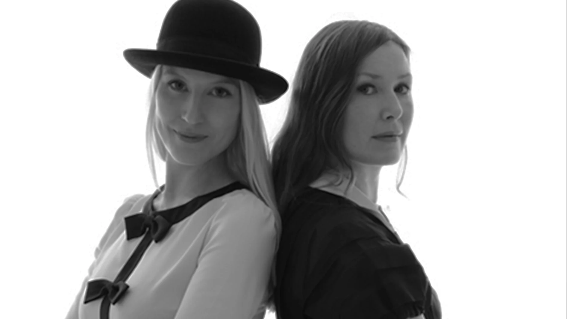 Svartvitt fotografi av två kvinnor som står rygg mot rygg. En bär hatt och blonda rosetter, den andra har långt mörkt hår.