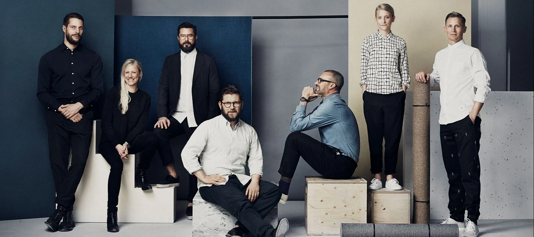 Sju personer, fem män och två kvinnor, i lediga, snygga kläder poserar i en modern studio med färgade bakgrunder och trälådor.
