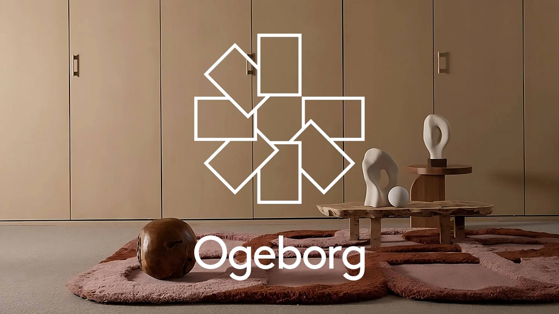 Minimalistiskt rum med ljusbruna skåp, rosa/brun matta, organiska träbord med vita skulpturer och träsfär. Ogeborg-logga.