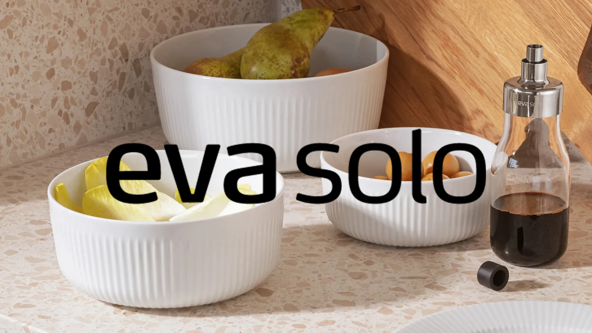 Vita räfflade skålar med päron och endive, samt en glasflaska med dressing på en spräcklig bänk. Varumärkestext 'eva solo'.
