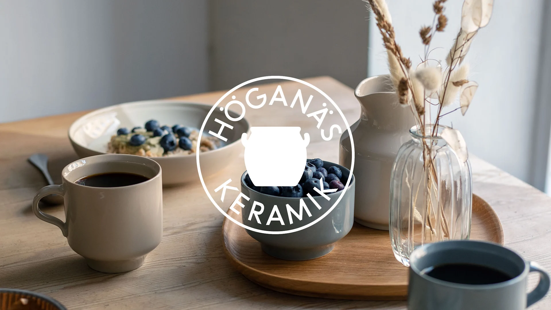 Frukostbord med kaffemuggar, skålar med blåbär och havregrynsgröt, kanna och vas, med Höganäs Keramik-logotyp.