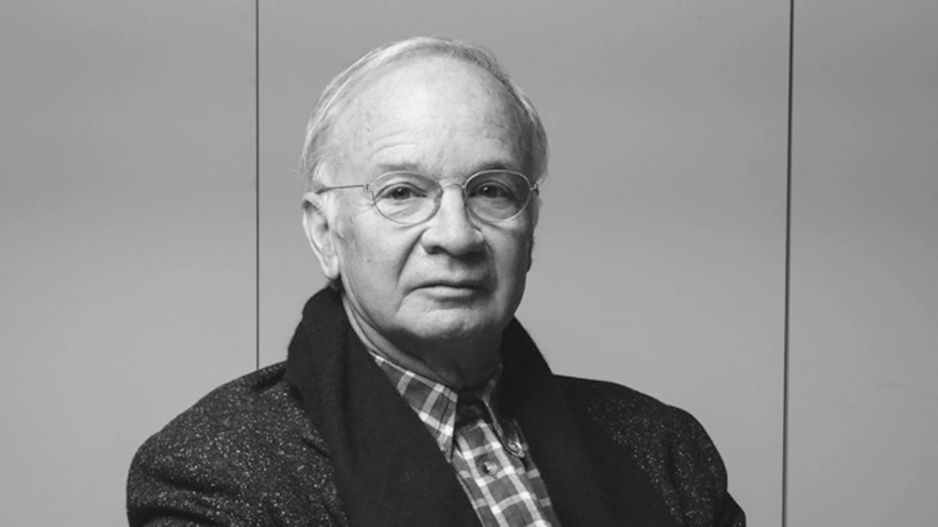 Svartvitt porträtt av en äldre man med glasögon, iklädd rutig skjorta och mörk jacka.