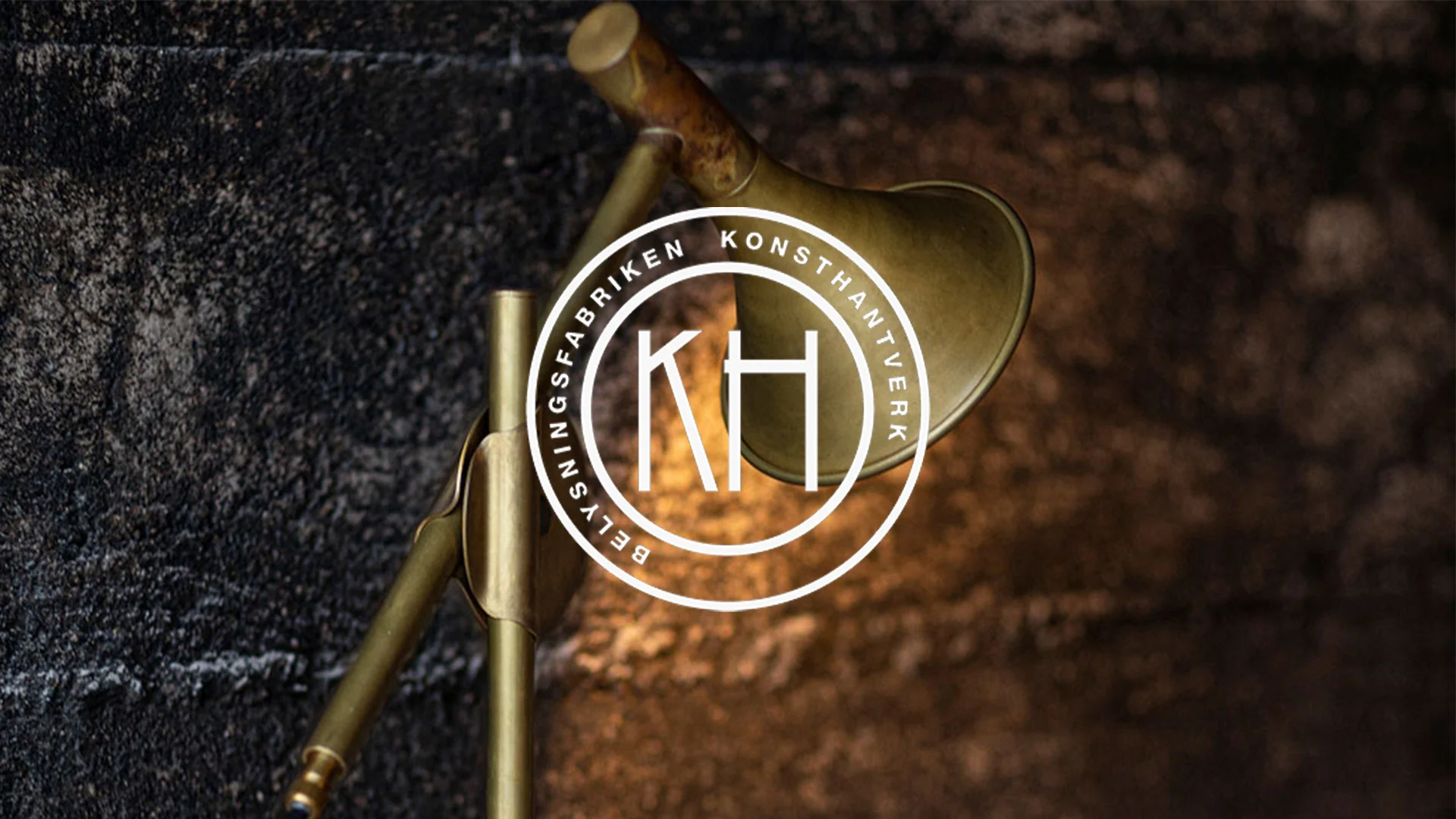 Mässingsfärgad lampa mot en strukturerad mörk vägg, med en vit cirkulär logotyp som visar 'KH' och texten 'Belysningsfabriken Konsthantverk'.