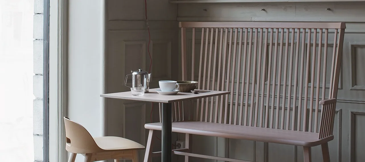 Fyrkantigt bord med tekanna och kopp, beige stol och rosa bänk mot en panelvägg.