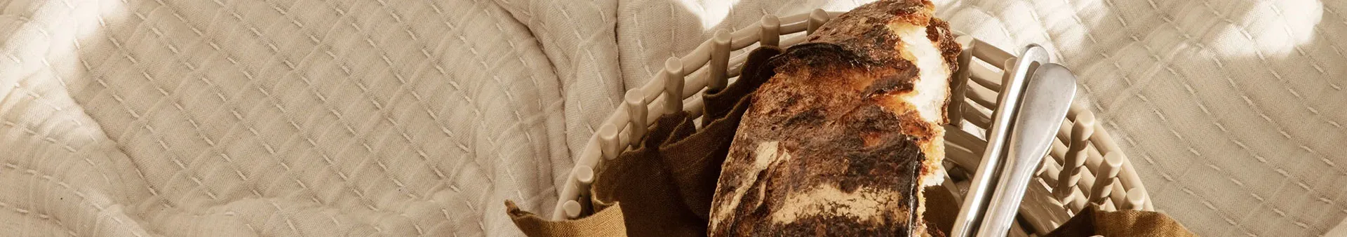 Ett rustikt bröd i en ljus, vävd korg med silvertång, placerat på en quiltad gräddfärgad filt.