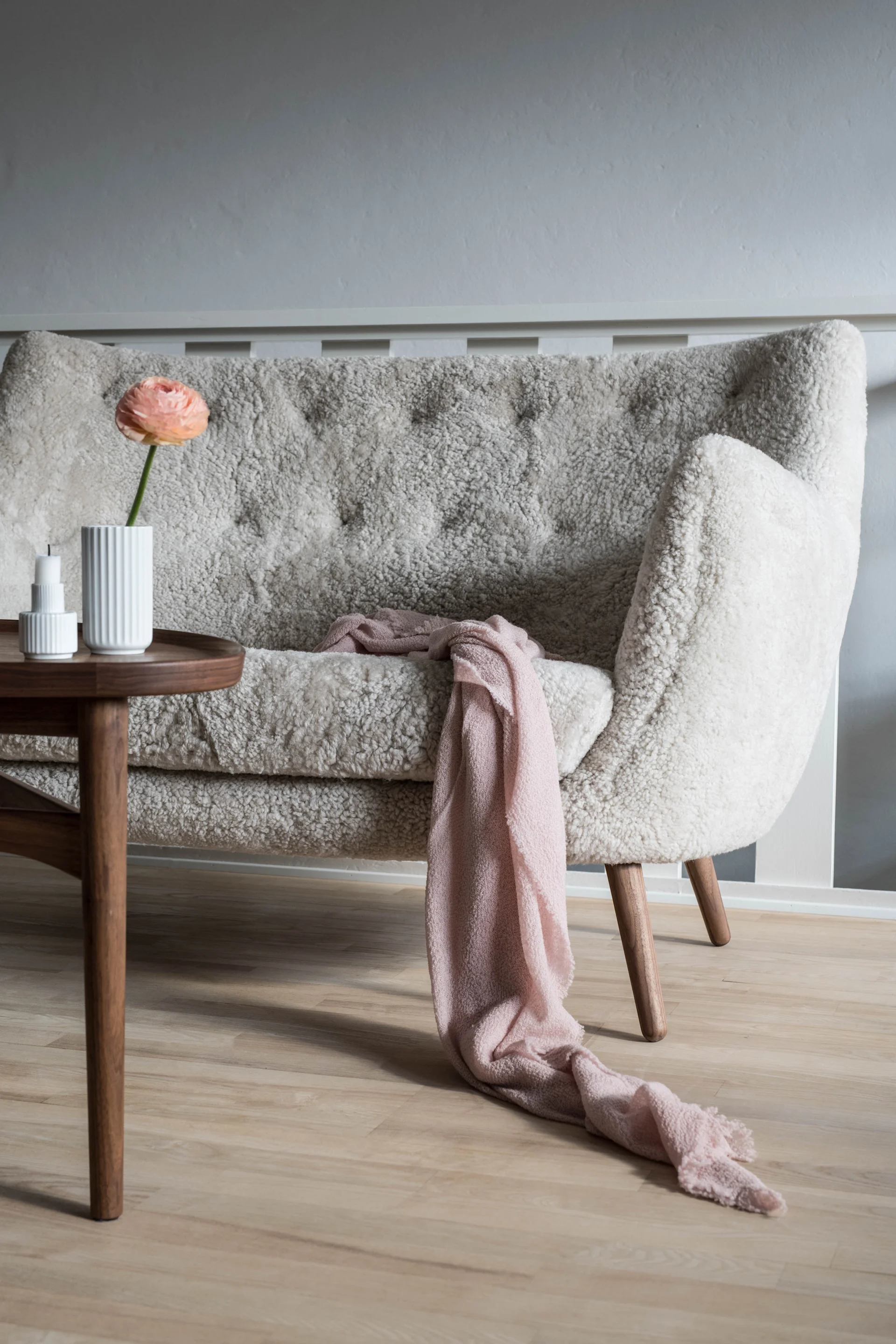 En ljus, fluffig soffa med en rosa filt, en persikofärgad blomma i en vit vas och en vit ljusstake på ett brunt sidobord.