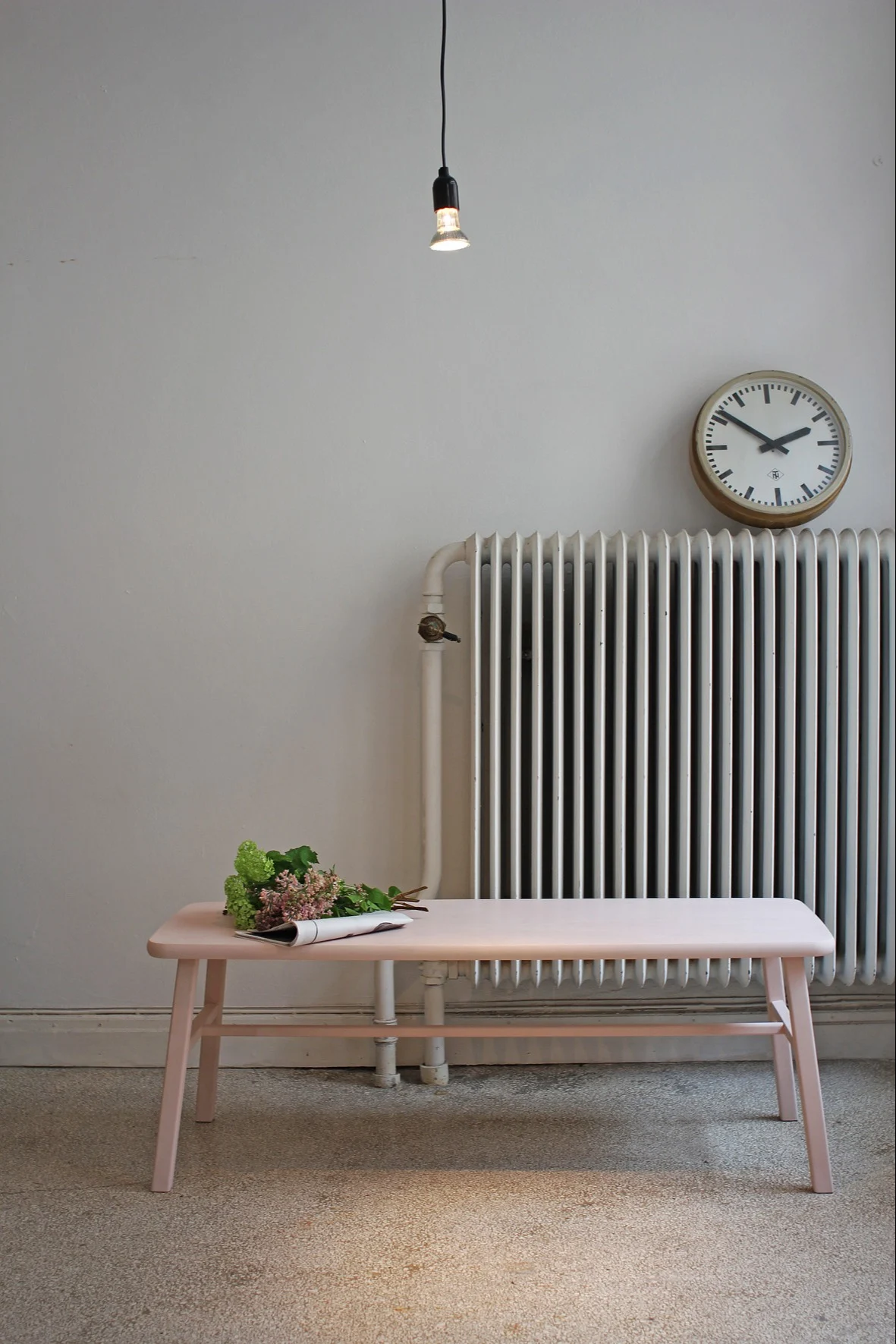 Ljusrosa bänk med blommor och tidning, vit radiator och rund väggklocka.