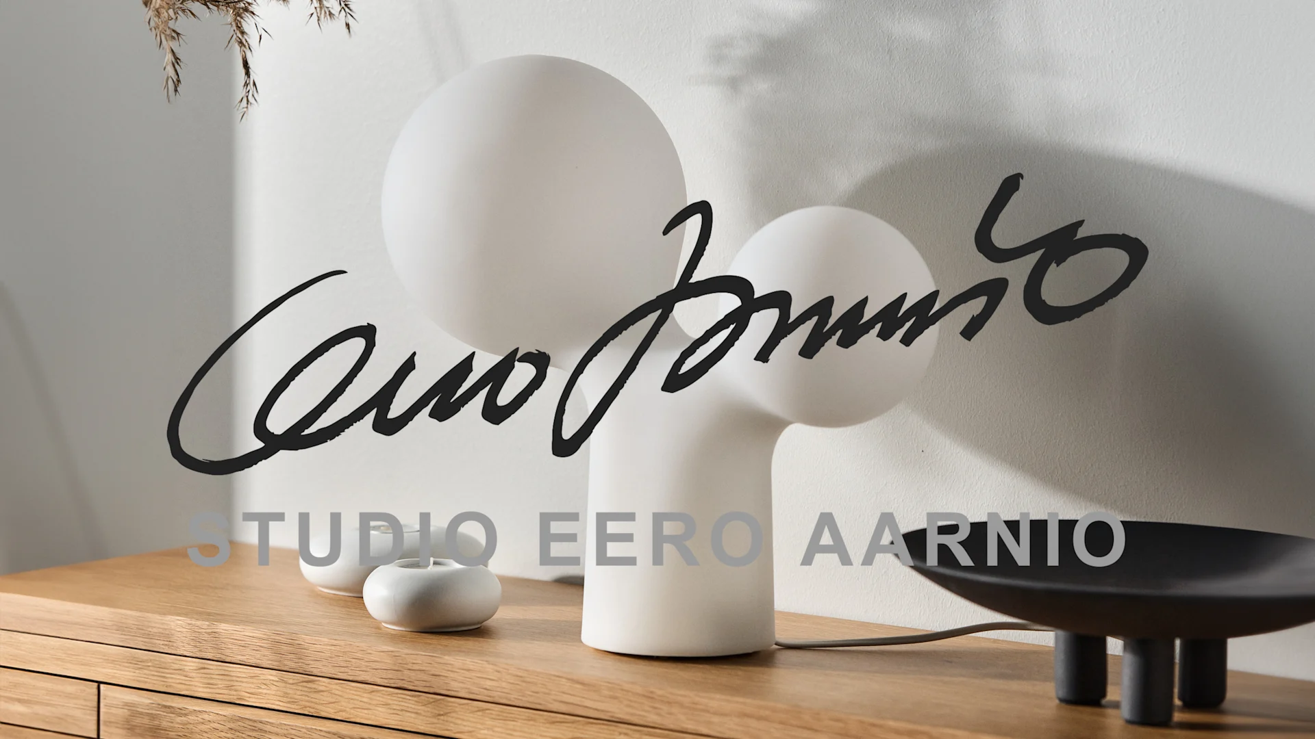 Vit skulptural bordslampa med sfärer, små vita ljushållare och en svart skål på en träyta. Text: STUDIO EERO AARNIO.