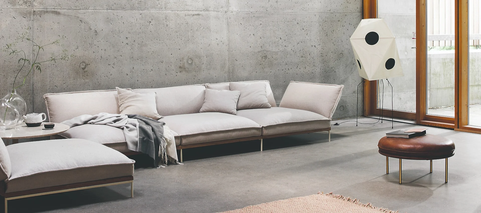 Ljusbeige modulsoffa med kuddar och en pläd i ett rum med betongvägg och stora fönster. En brun fotpall och golvlampa.