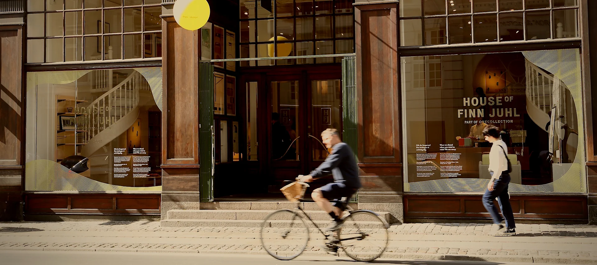 Gatuvy framför en butik med stora fönster som visar möbler, en cyklist och en person som går på trottoaren.