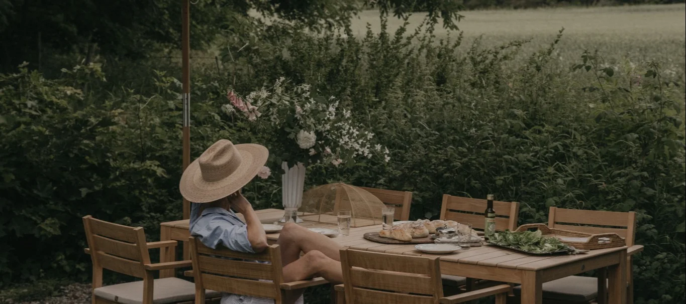 En person med stråhatt sitter vid ett utomhus matbord i trä med bröd, sallad och blommor.