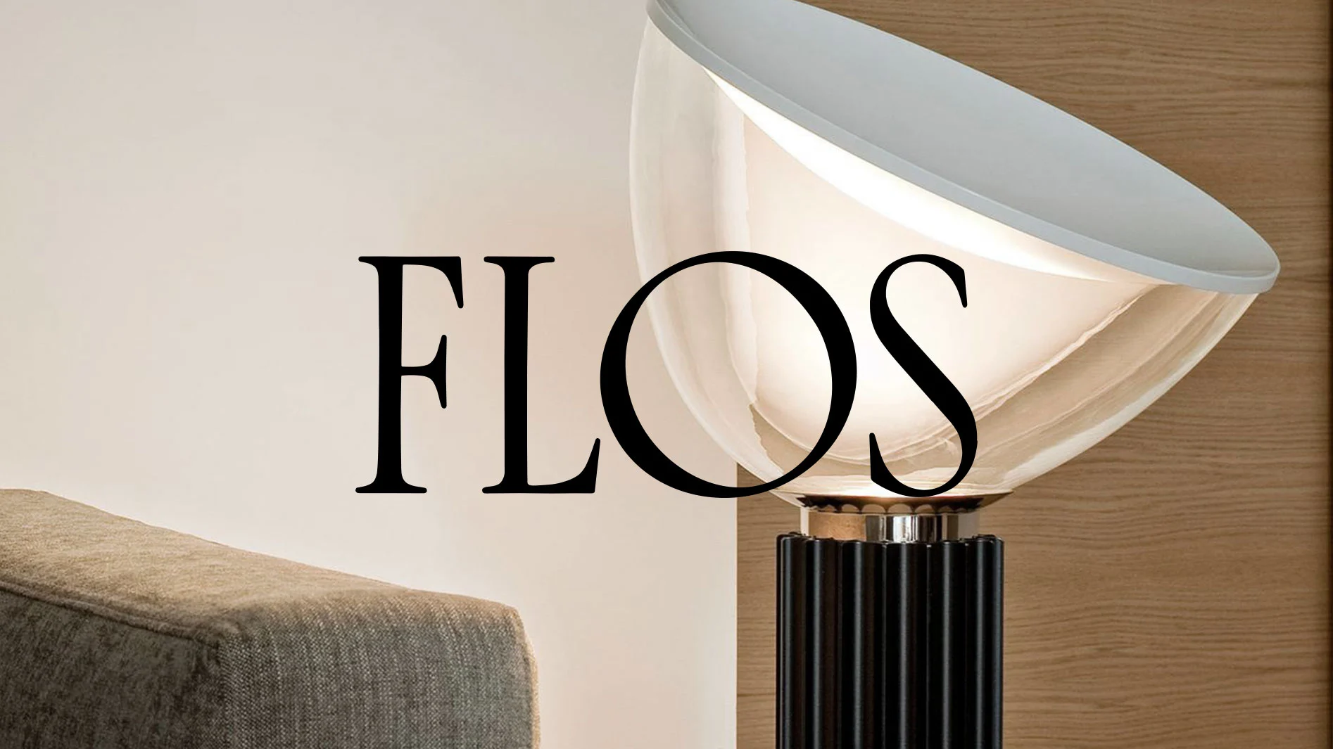 Svart text FLOS över en modern lampa med klar och vit skärm, mörk räfflad fot, och en grå soffa.