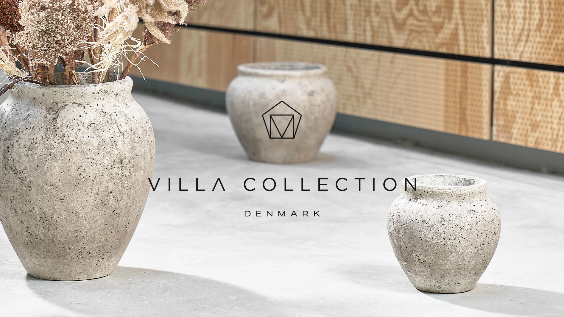 Tre rustika betongvaser, en fylld med torkade blommor. Märket "VILLA COLLECTION DENMARK" är synligt.