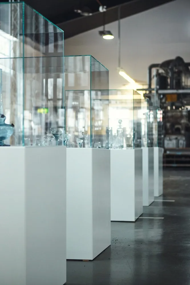 En rad vita piedestaler med glasvitriner som visar glasföremål i ett modernt utställningsrum.