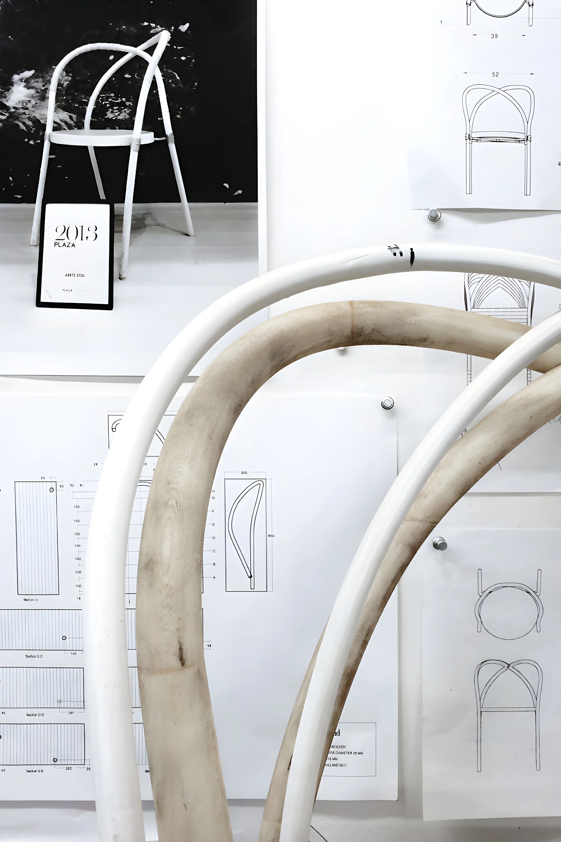 Stoldesigner utställda: en vit prototypstol, böjda ramdelar och stolritningar med en '2013 PLAZA'-skylt.