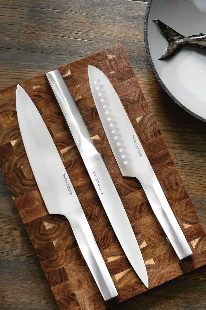 Tre silverfärgade knivar med olika blad ligger på en rutig skärbräda i trä. En fisksvans syns på en tallrik.