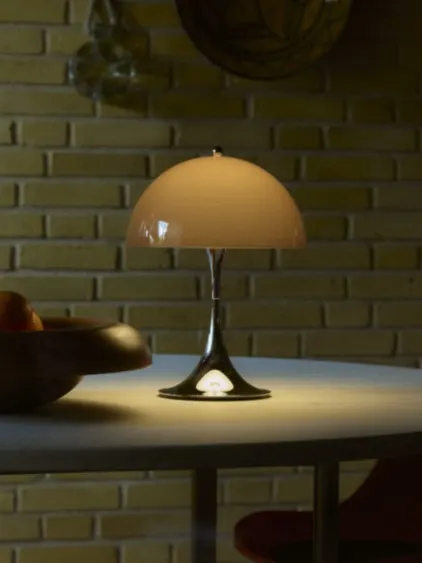 Bordslampa med beige svampformad skärm och glänsande metallfot, på ett bord mot en tegelvägg.