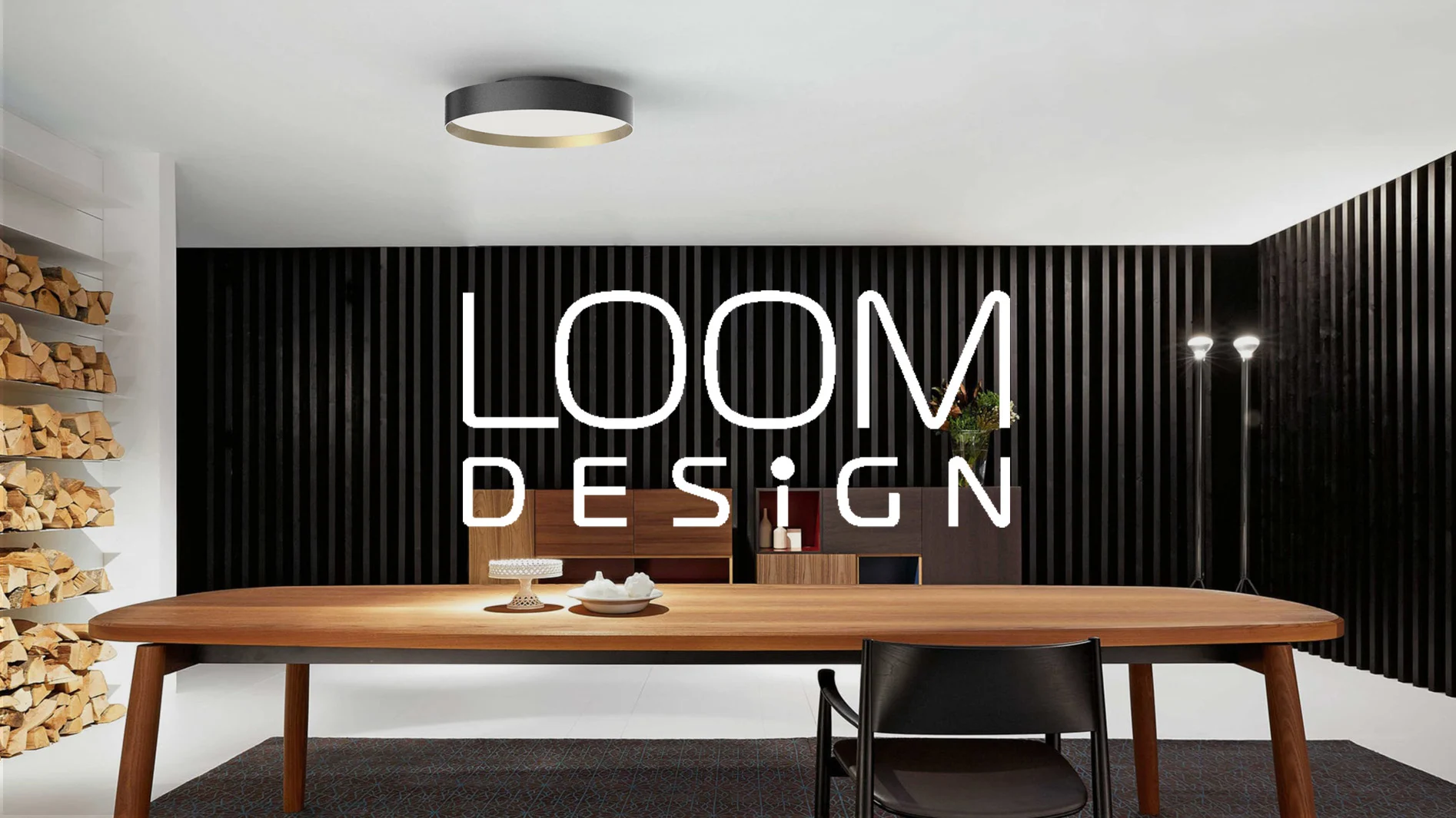Ett modernt rum med ett stort matbord i trä, svart stol, en hylla med vedträn och en svart ribbvägg med texten "LOOM DESIGN".