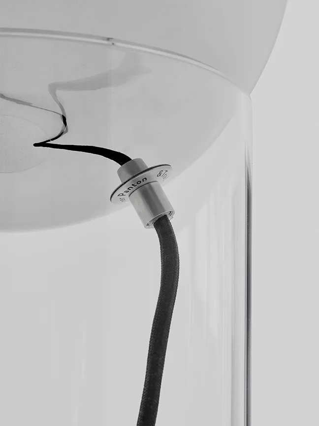 Närbild av en lampas upphängning med en svart flätad kabel, ett metallfäste med texten "Panton" och en genomskinlig eller frostad glasskärm.