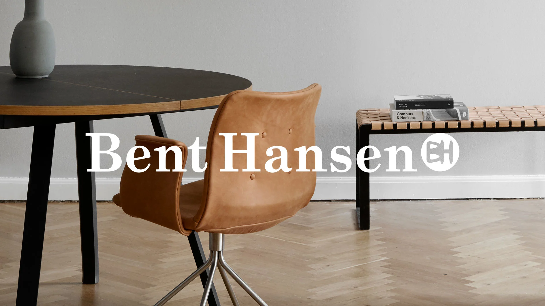 Logotypen Bent Hansen över ett runt matbord, en brun läderstol och en flätad bänk i ett modernt rum.