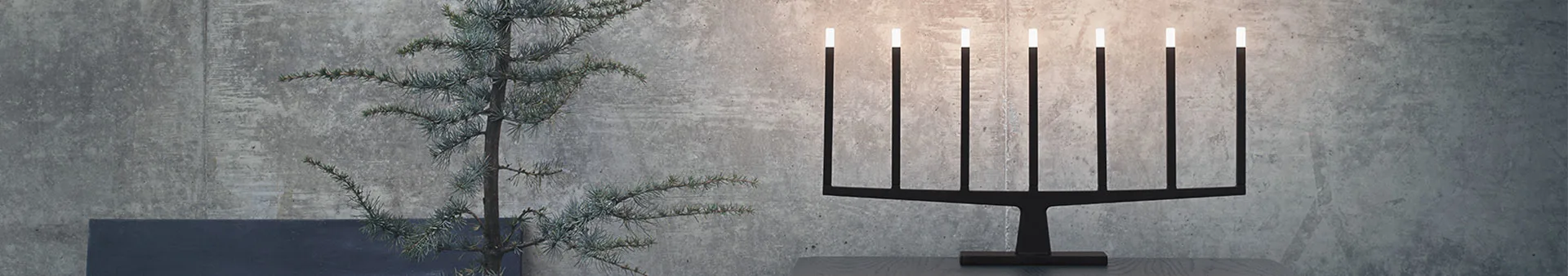 Svart, minimalistisk ljusstake med sex lampor, och grön granris mot grå vägg.