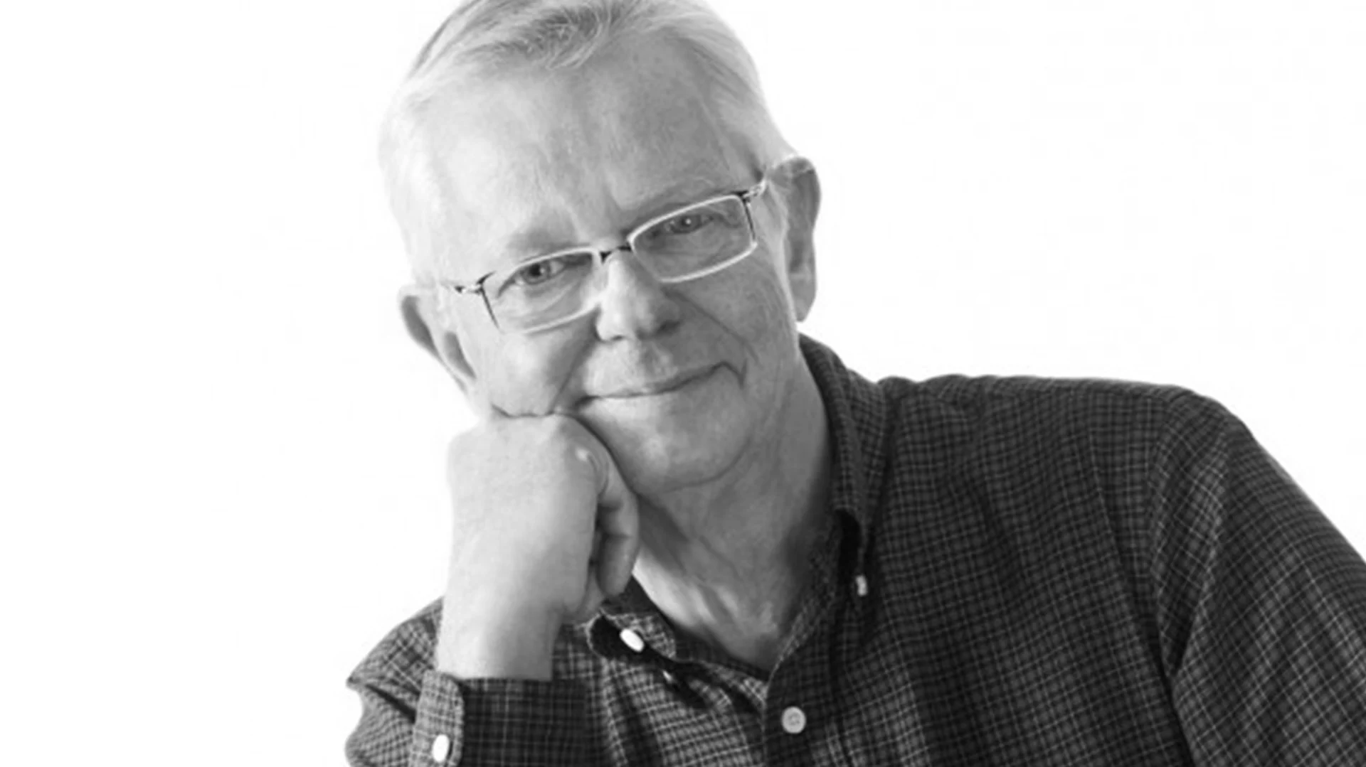 Svartvitt porträtt av en äldre man med glasögon och rutig skjorta, som vilar hakan i handen och ler.