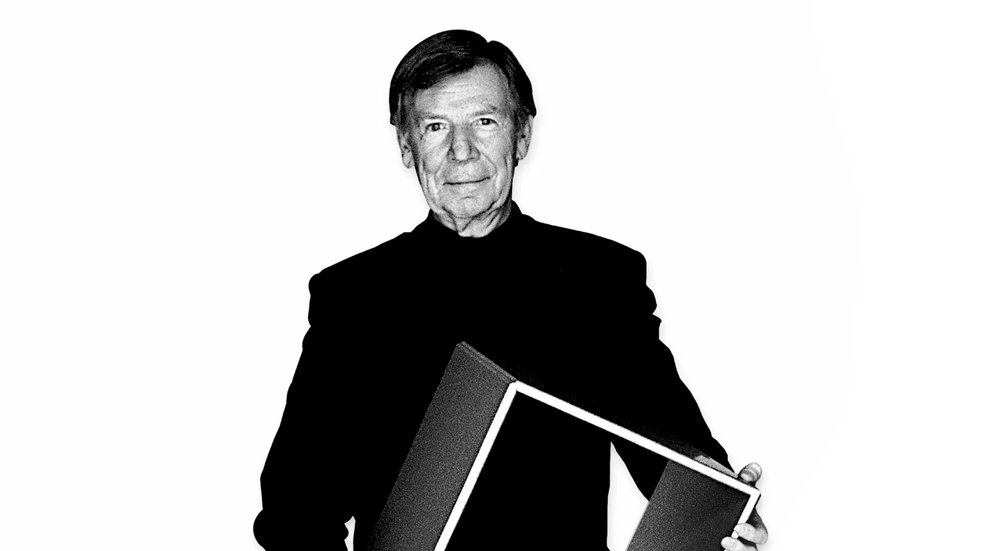 Svartvitt porträtt av en äldre man i mörk jacka, som håller ett geometriskt objekt.