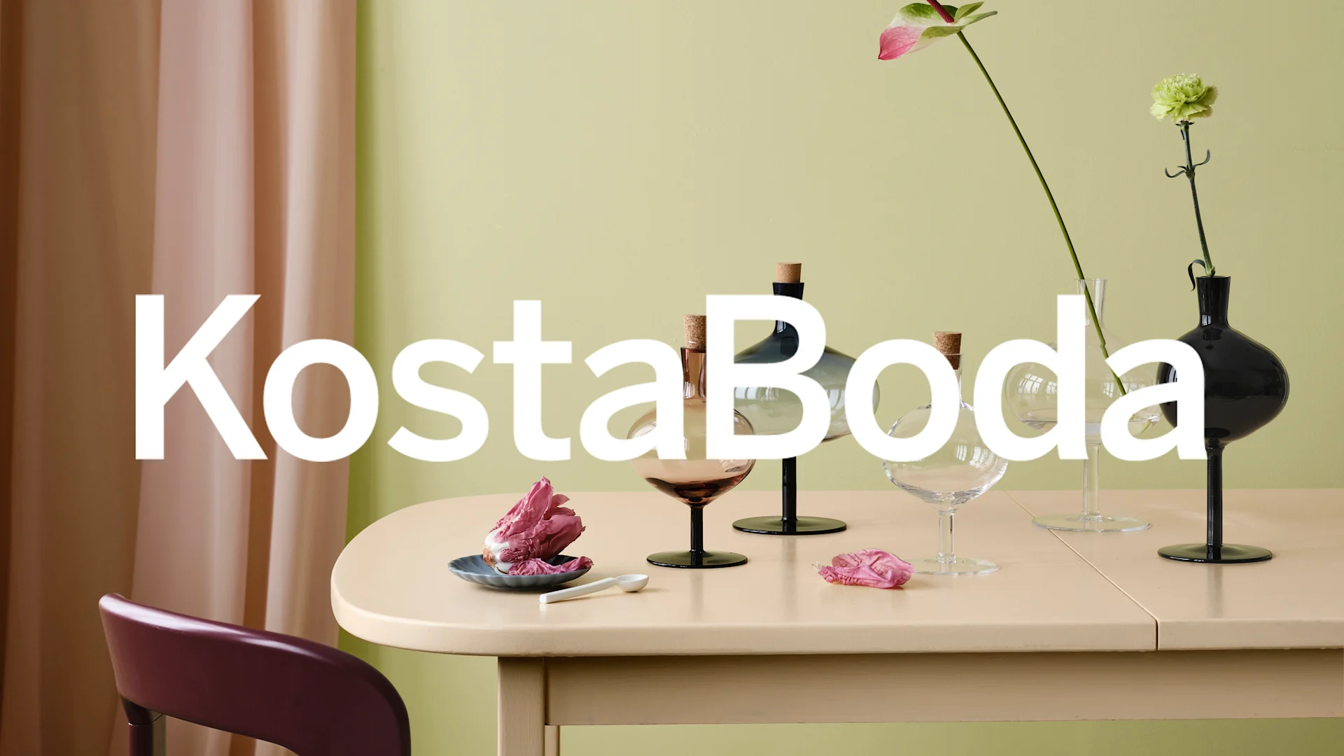 KostaBoda text över ett beige bord med glasflaskor, vaser, blommor och ett rött kålblad.