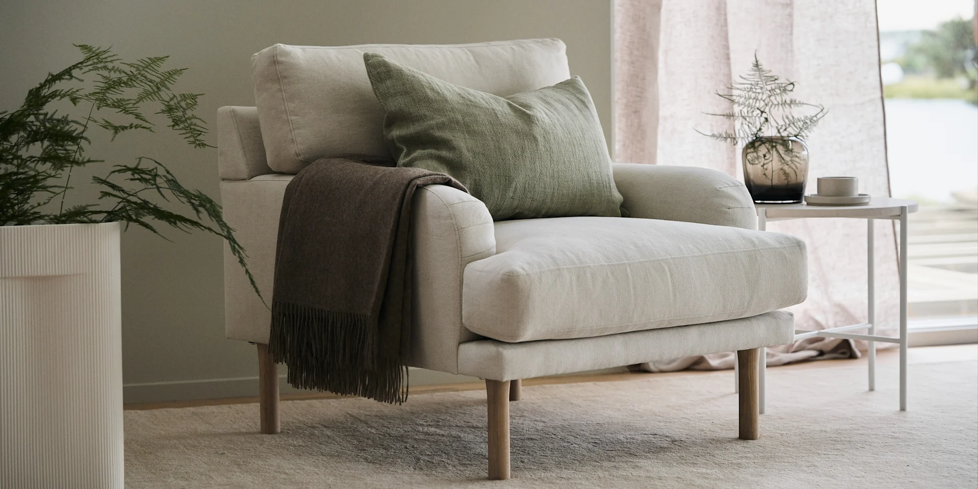 Lidnäs fåtölj i beige från varumärket 1898 placerad i vardagsrum på beige ryamatta med det runda lilla kalkstensbordet Aplaryd bredvid. Fåtöljen är försedd med gröna kuddar och grön pläd.