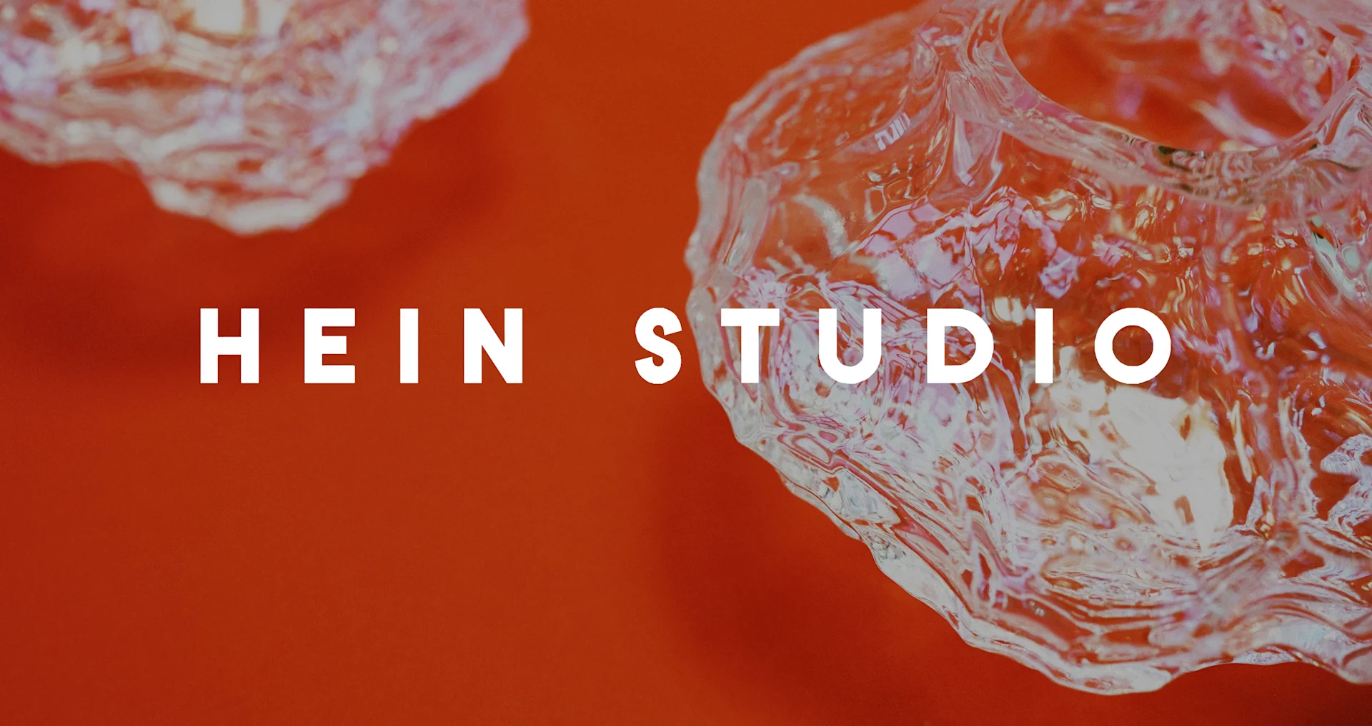 Vit text 'HEIN STUDIO' på orangeröd bakgrund, flankerad av två genomskinliga, strukturerade glasobjekt.