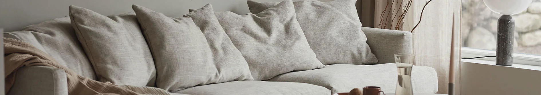 Ljus soffa med kuddar och en beige pläd, vid ett fönster med en marmorlampa och glaskaraff.