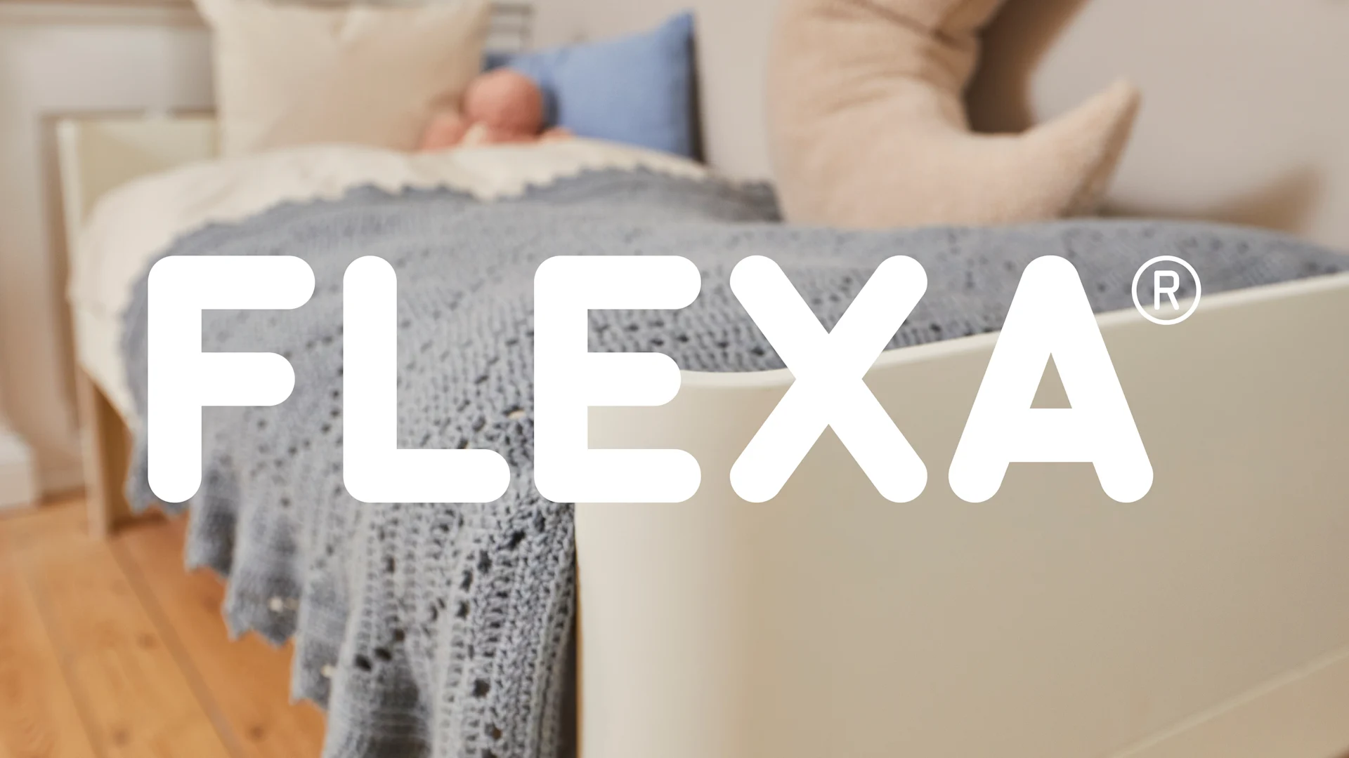 Säng med blå stickad filt, ljusa kuddar och vit FLEXA-logotyp.