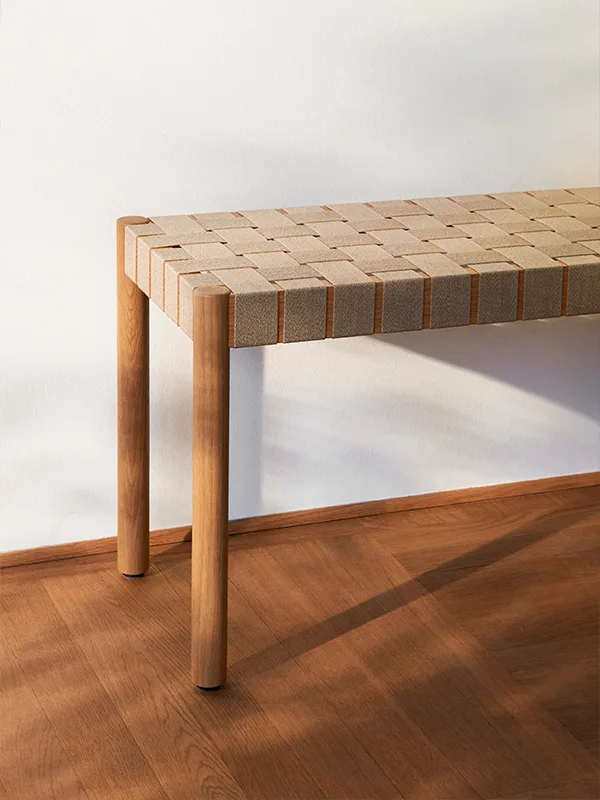 En träbänk med vävd sits i neutrala färger står mot en vit vägg på ett trägolv.