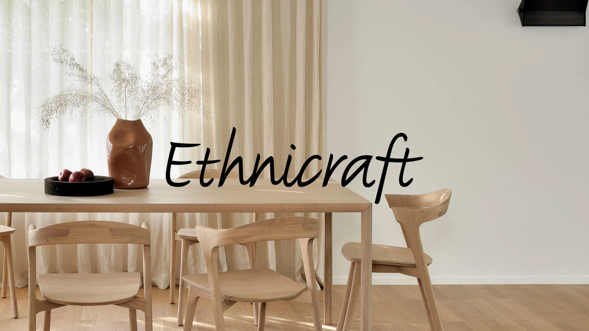 Matsal med ljust träbord och stolar, vas med torkat gräs, frukt och Ethnicraft-logga.