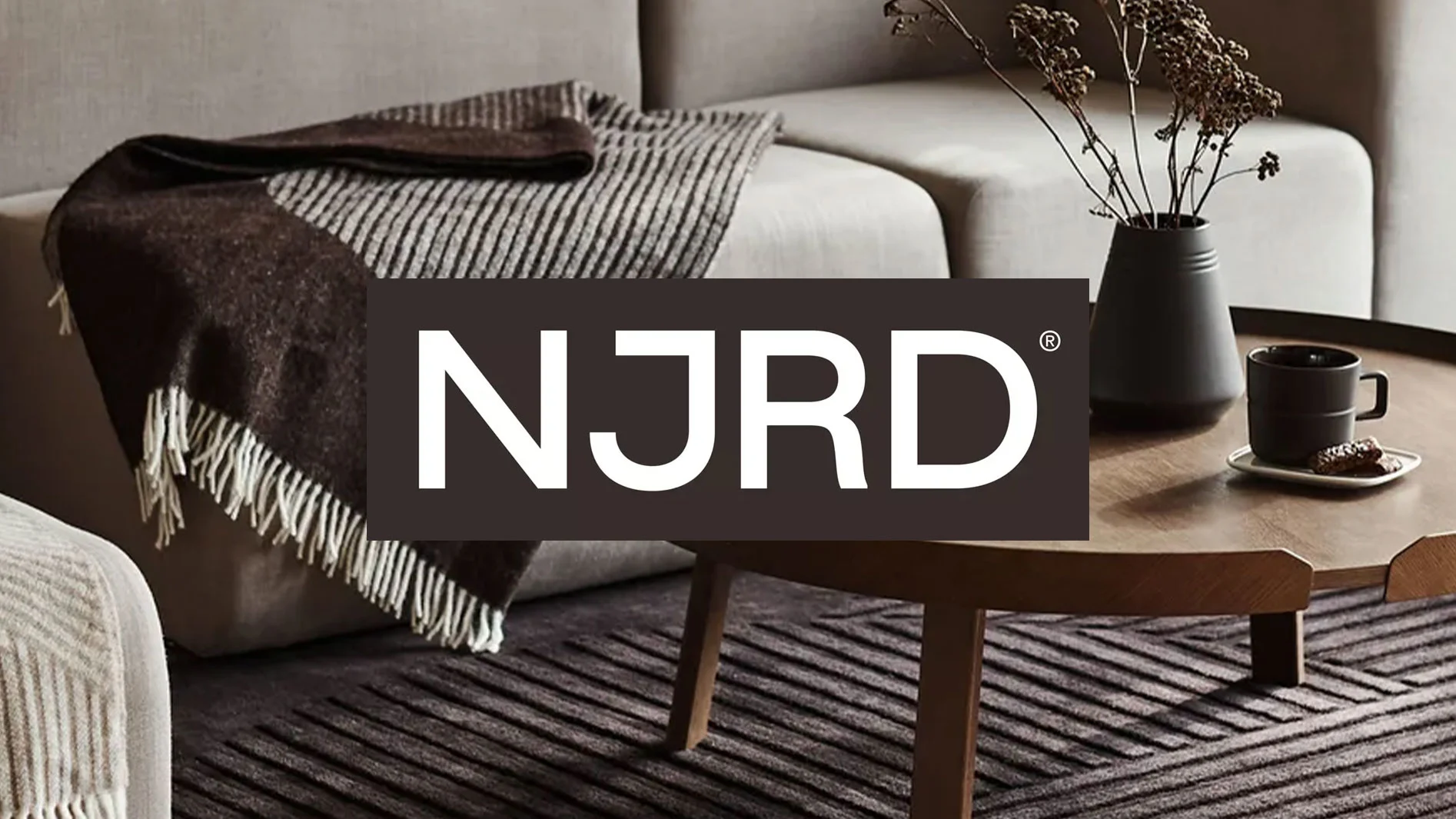 "NJRD" logotyp över en mysig vardagsrumsinredning med brun filt på soffa, soffbord med vas och kopp, och mörk, mönstrad matta.