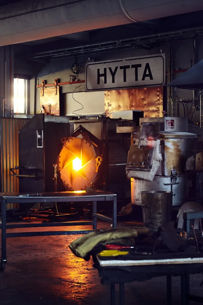 Interiör av en glasblåsarverkstad med en glödande smältugn och skylten «HYTTA».