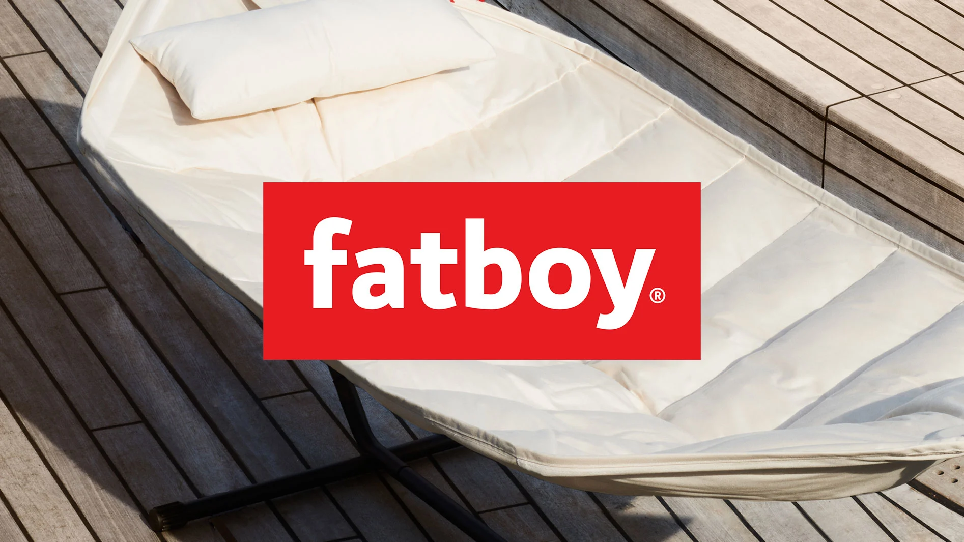 Gräddvit utomhus solsäng med kudde på ett trädäck, med röd Fatboy-logotyp överlagd.
