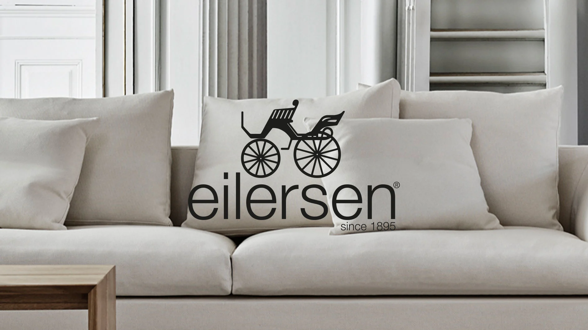 Ljusbeige soffa med kuddar. En kudde har en svart logotyp med en hästdragen vagn och texten "eilersen since 1895".
