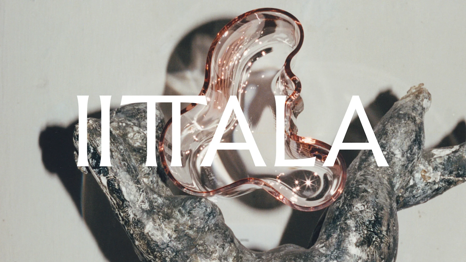 IITTALA-text över ett vågigt, bärnstensfärgat glasobjekt som vilar på en mörk, texturerad yta.